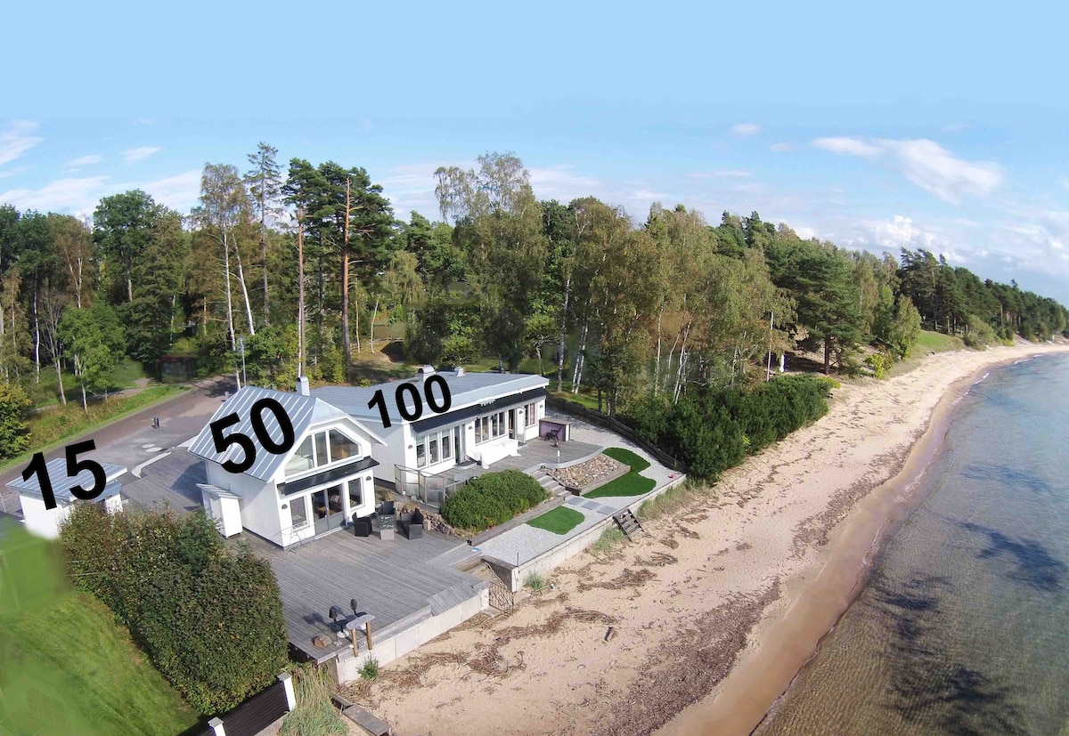 An aerial view captures a contemporary home situated along a sandy beach, framed by lush green trees. The large deck extends towards the water, providing direct access to the beach. The serene landscape features a gentle shoreline and tranquil waters.