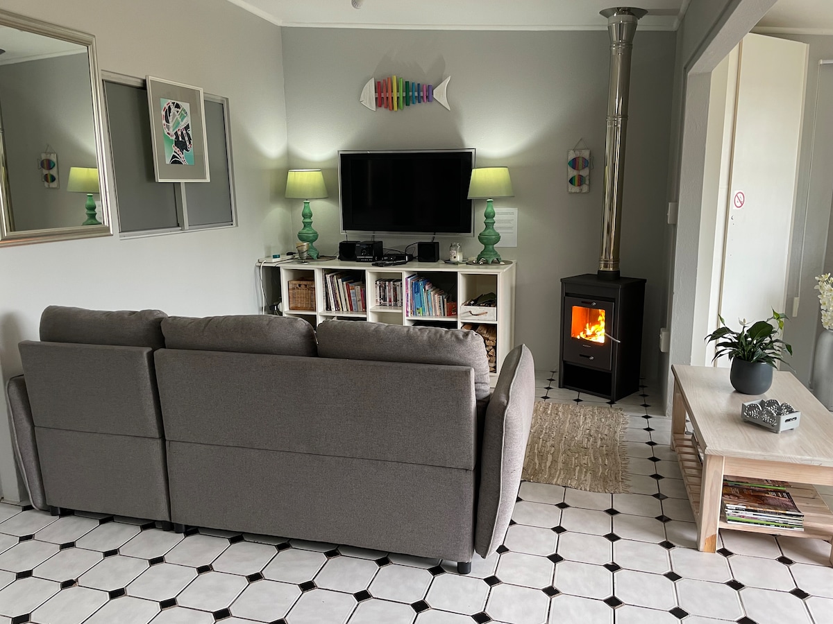 A comfortable living area features a grey sofa positioned towards a wall-mounted flat-screen TV. A log burner emits a warm glow, while shelves hold a selection of books and DVDs. Soft lighting from green lamps and decorative mirrors add to the inviting ambiance.