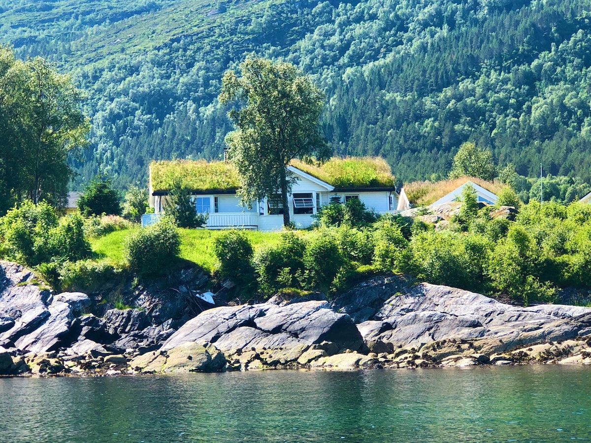 A house with a grass-covered roof is positioned on a gentle slope overlooking the water. Lush greenery and trees surround the property, blending naturally with the mountainous backdrop. Rocks are visible at the water's edge, contributing to the serene coastal environment.