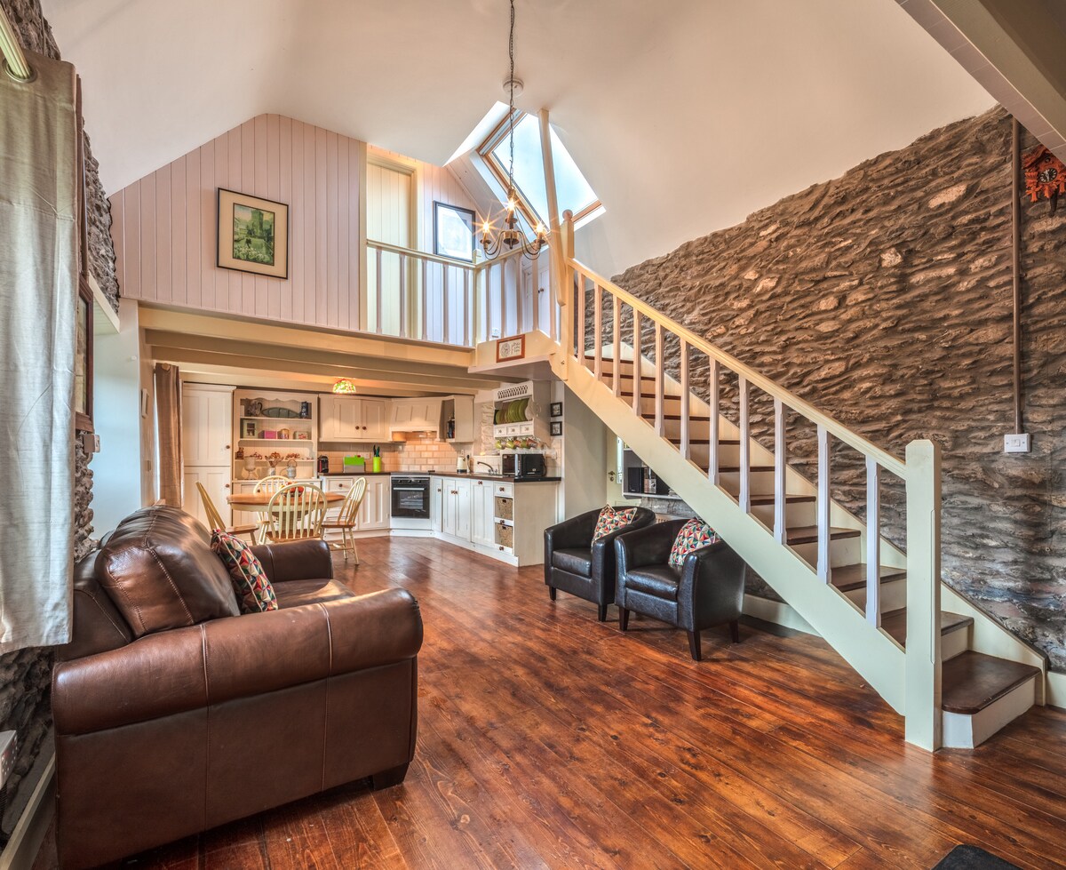 The open-plan living area features a high ceiling with skylights allowing natural light to fill the space. A staircase leads to the upper level. Two leather sofas are positioned on a wooden floor, and a dining area is visible in the background near the kitchen.