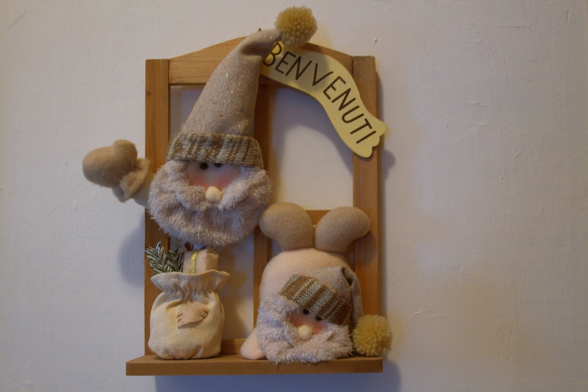 A small wooden frame is mounted on the wall, featuring plush decorative figures. Two characters, one resembling a gnome and the other a bunny, wear soft, textured hats and scarves. A sign reading 'Benvenuti' is positioned above them, enhancing the welcoming touch of the display.