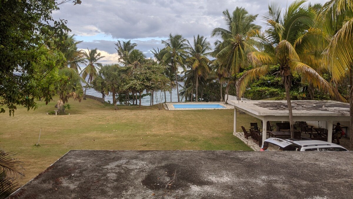 A wide view captures a grassy area leading to a pool, surrounded by palm trees and ocean in the background. The pool is framed by a simple patio area with seating. The overall setting conveys a sense of privacy and tranquility.