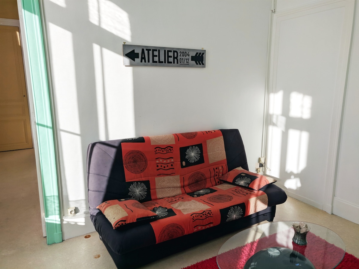 A comfortable sofa with an orange and black patterned throw is located in a bright space. A glass coffee table sits in front of the sofa, reflecting the light streaming in through nearby windows, which feature soft, natural light and distinct shadows.