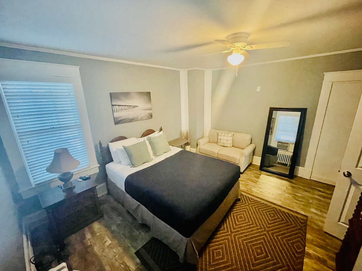 A cozy bedroom features a queen-size bed adorned with soft linens. A ceiling fan adds comfort, while a large mirror and a sectional sofa provide additional space. Natural light streams through the window, highlighting the warm wood tones and patterned area rug.