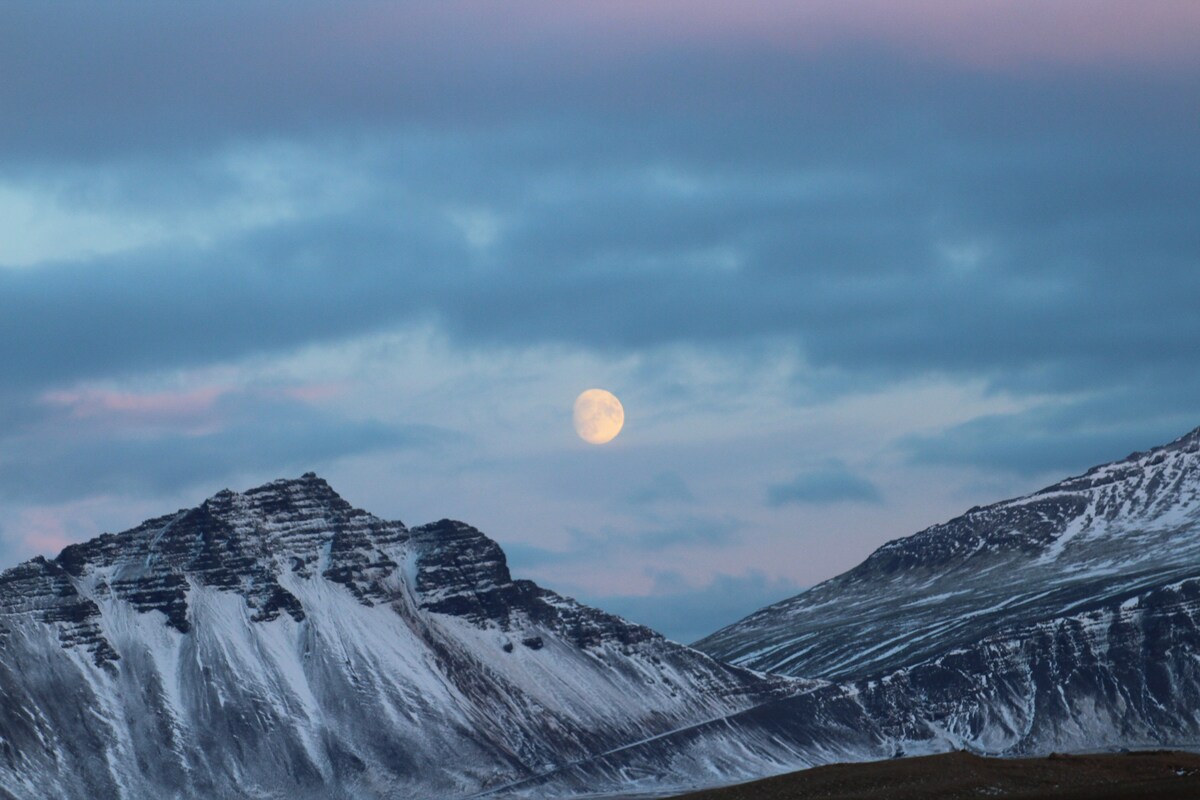 A large full moon is positioned in the sky above rugged snow-capped mountains. The landscape is accented by soft pastel hues in the clouds, creating a serene twilight atmosphere. The terrain displays a mixture of rocky outcrops and snow-covered slopes.