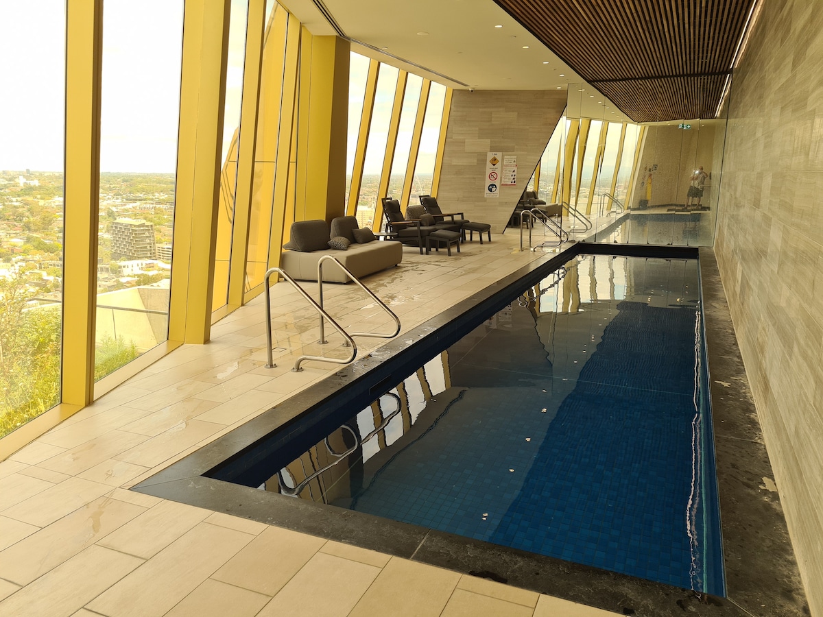 A sleek indoor swimming pool features large glass windows that offer panoramic views of the surrounding area. Sun loungers are positioned alongside the pool, while the light-colored stone flooring and wooden ceiling create a contemporary and spacious atmosphere.