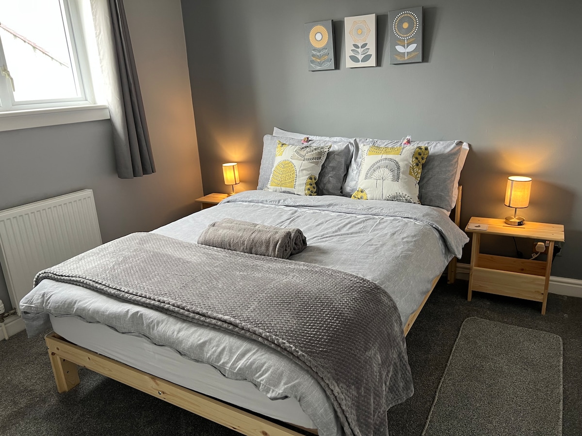 A spacious bedroom is furnished with a wooden bed frame covered in soft gray bedding. Two bedside tables hold warm lamps, and decorative art prints are displayed above. Light filters through a nearby window adorned with gray curtains, enhancing the serene atmosphere.
