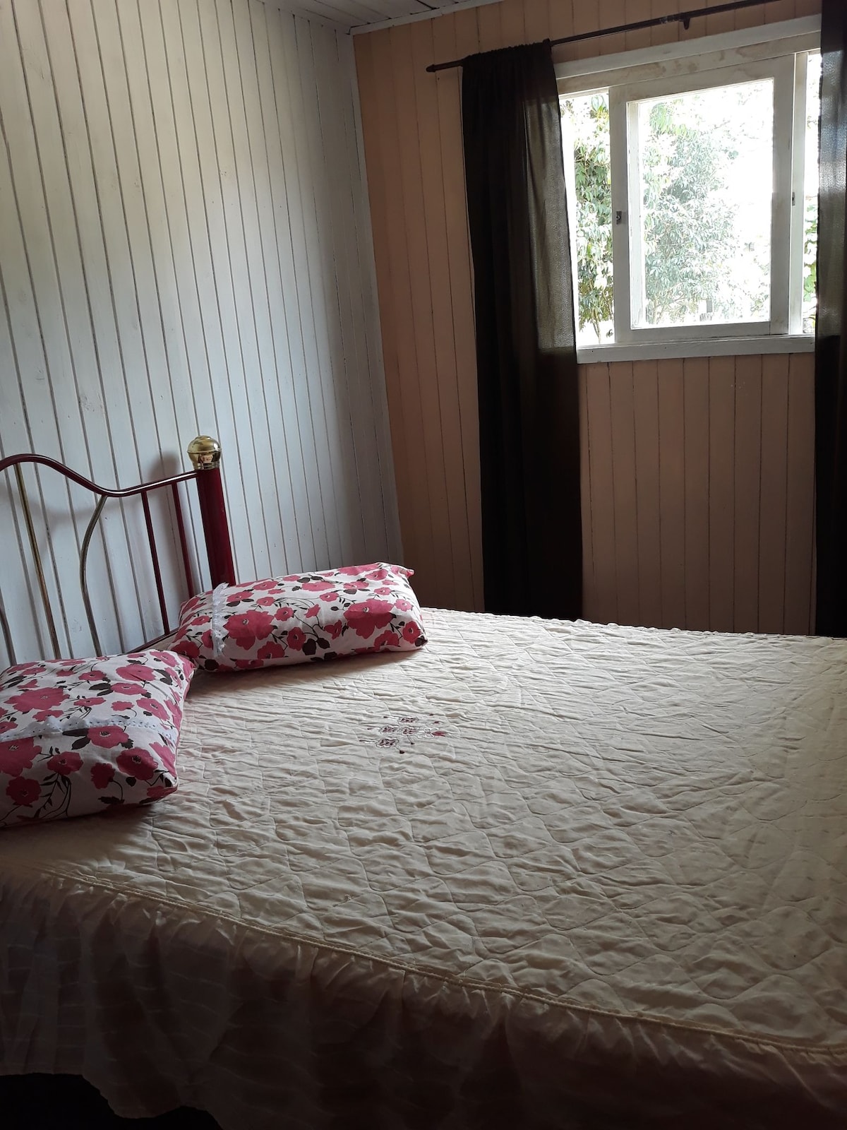A modest bedroom is presented, featuring a metal bed frame with a light-colored quilted coverlet. Decorative pillows with floral patterns are arranged on the bed. A window allows natural light to enter, framed by dark curtains that enhance privacy.