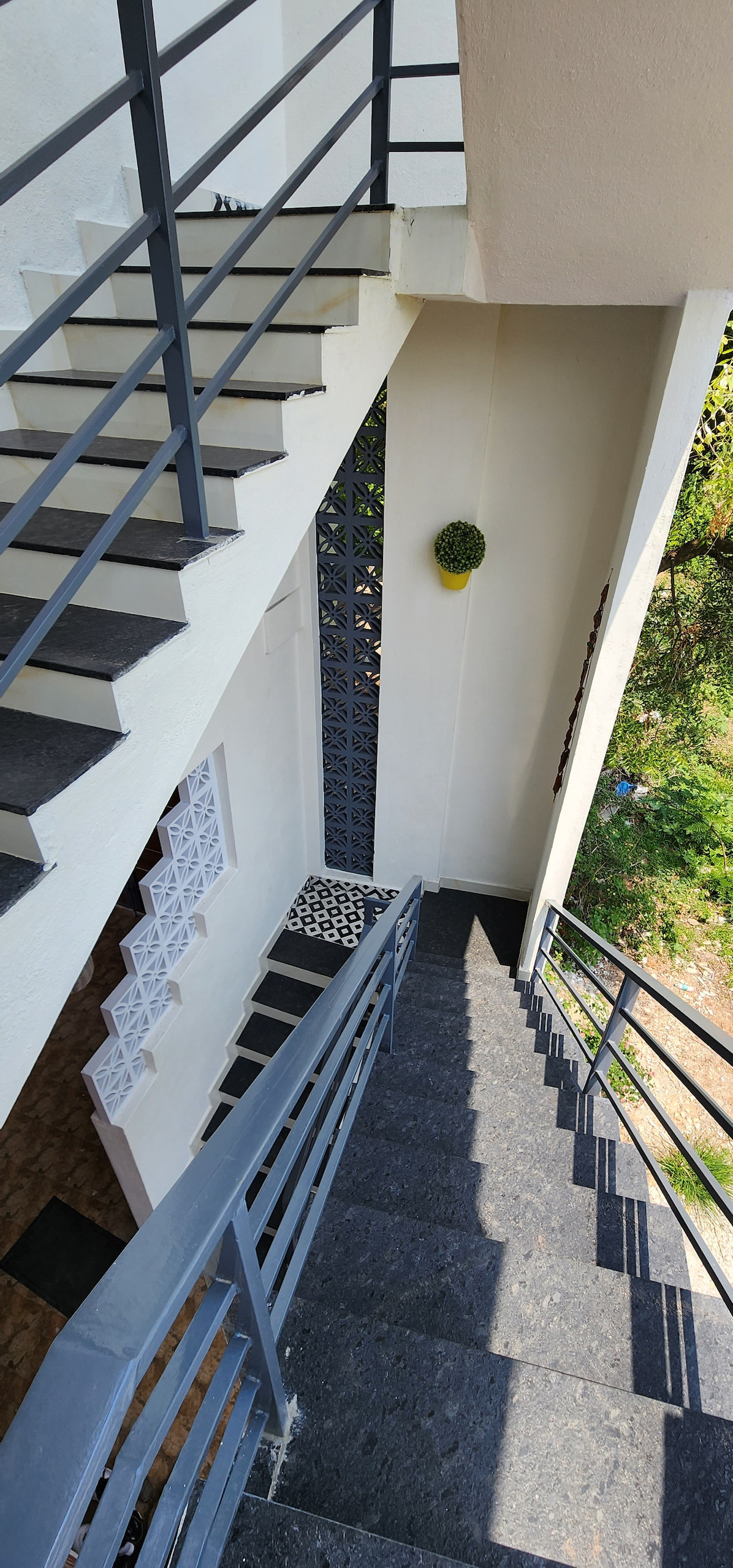 A staircase is shown with a modern railing in a duplex setting. Natural light filters through the nearby window, highlighting the textured stone steps. A small decorative plant is positioned on the wall, adding greenery to the clean, neutral walls.
