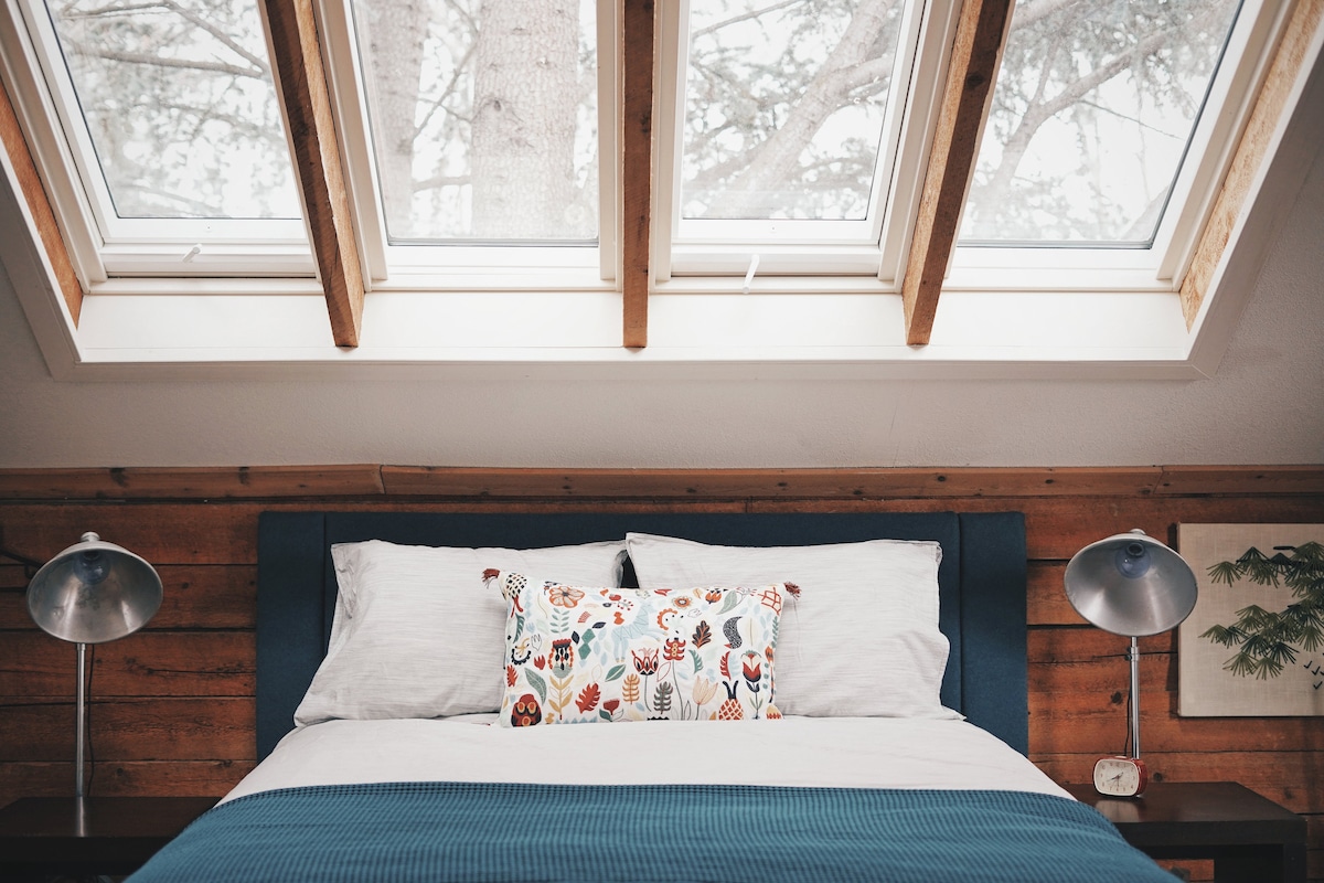 A spacious bedroom features a comfortable bed adorned with a decorative pillow. Natural light streams through skylights above, illuminating the wooden walls and creating a serene atmosphere. Two bedside lamps are placed symmetrically on either side of the bed.