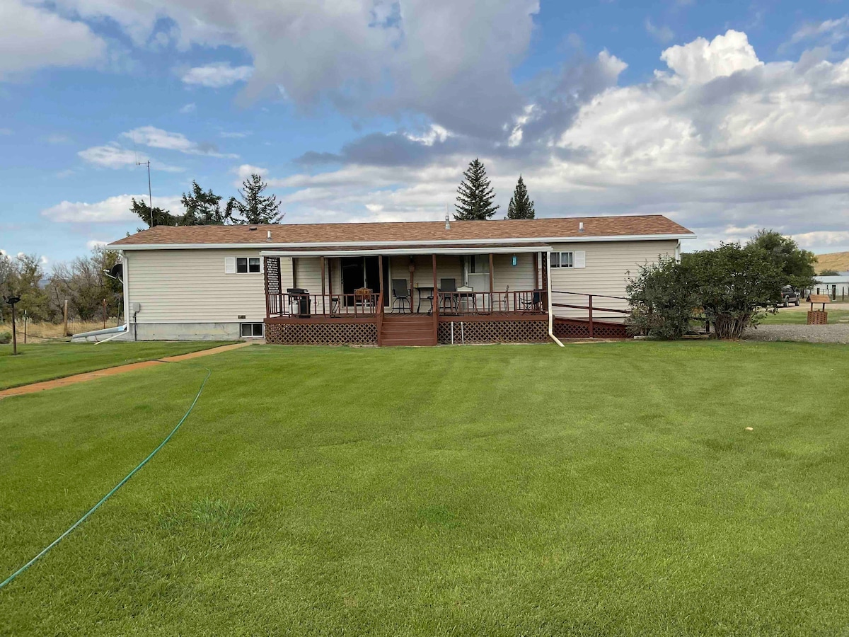 The exterior of a single-story home is displayed, featuring a covered deck with seating. Lush green grass surrounds the property, and a clear sky with scattered clouds creates an expansive backdrop. The structure is positioned against a backdrop of trees and open pastures.