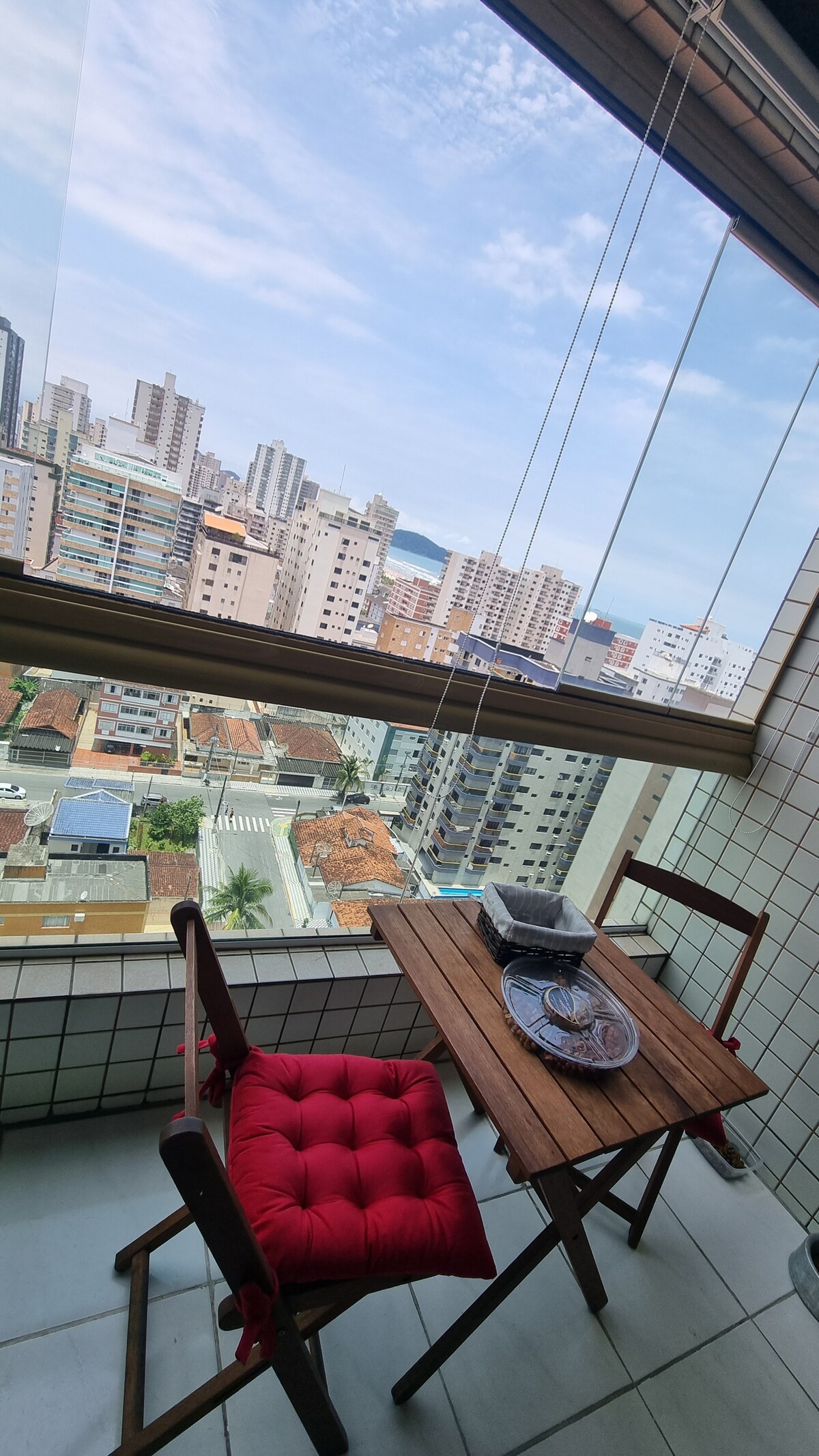 A small balcony features a wooden table accompanied by two chairs, one adorned with a red cushion. Large glass windows allow for a view of the cityscape and distant ocean, enhancing the outdoor experience.