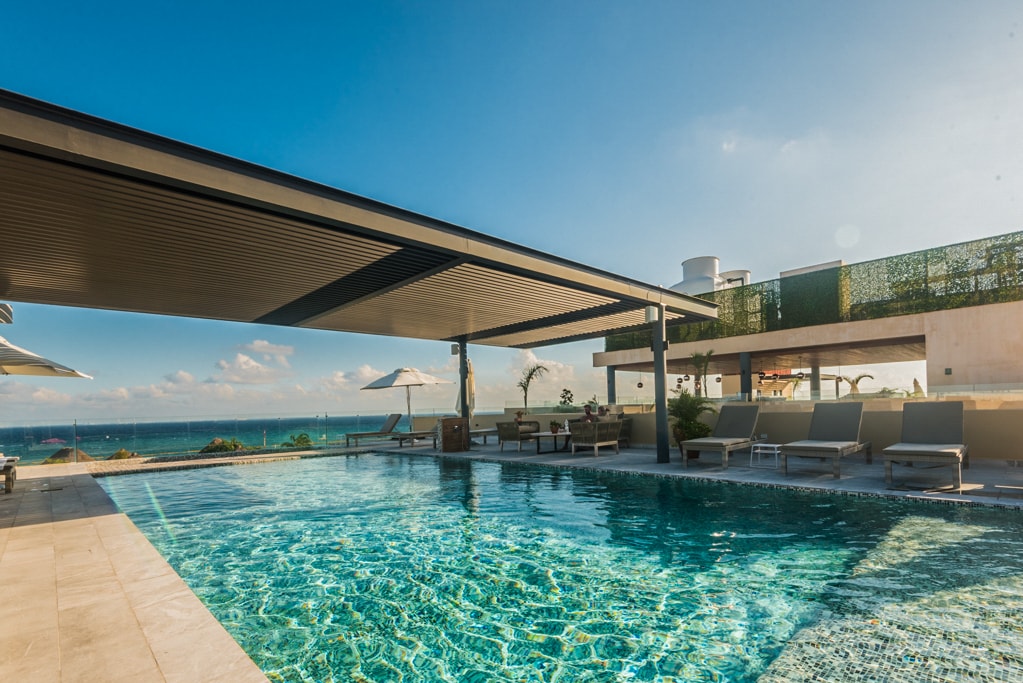 A rooftop pool area is featured, with clear blue waters reflecting the sky. Comfortable lounge chairs are arranged near the pool, and shaded spaces are provided by large umbrellas. The ocean is visible in the background, enhancing the serene atmosphere.