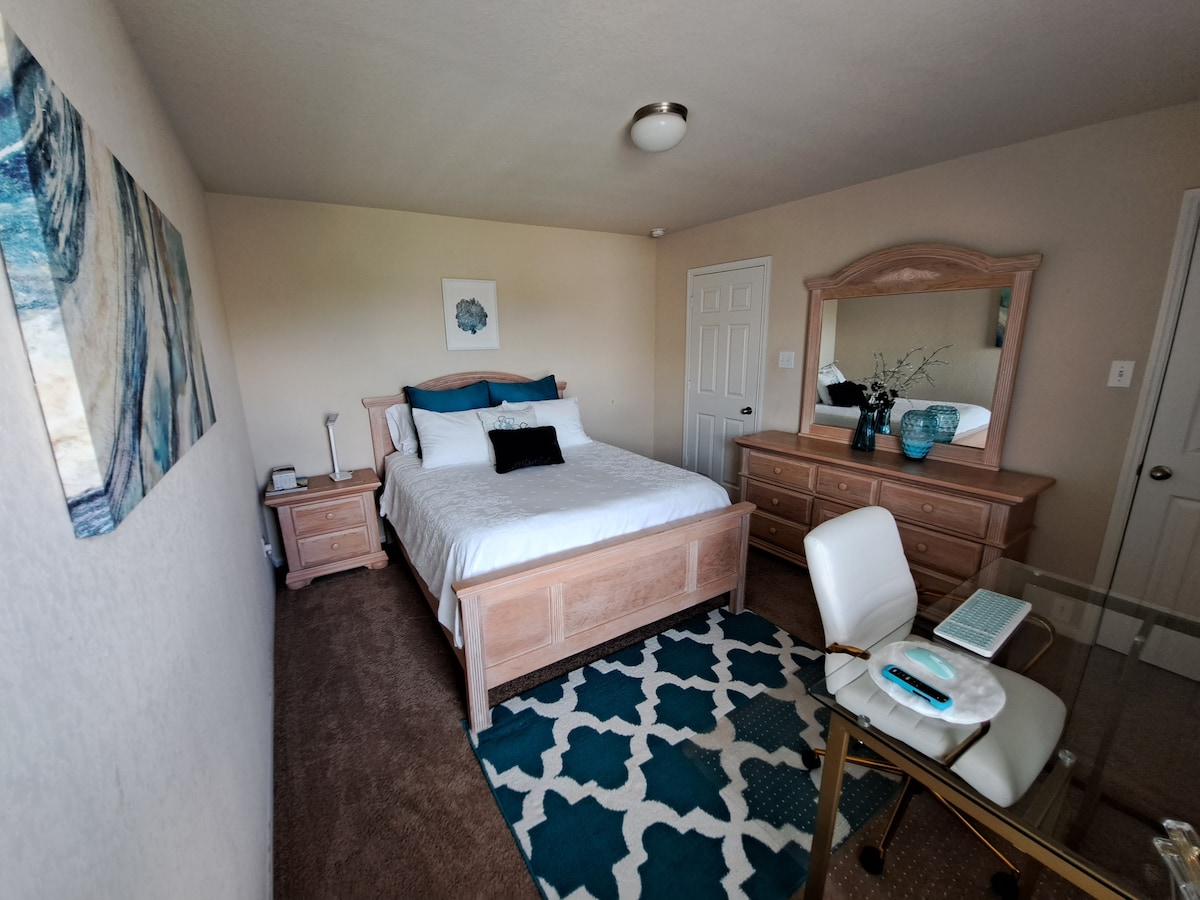 A spacious bedroom features a comfortable bed with crisp white linens and a decorative black accent pillow. A wooden dresser and mirror complement the light decor, while a decorative rug adds a pop of color. A desk is positioned near the window for workspace convenience.