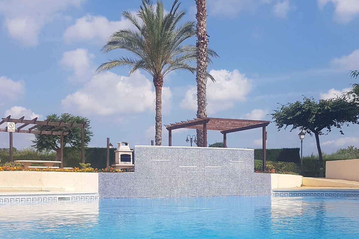 The image captures a serene pool area featuring clear blue waters and a decorative water feature. Lush palm trees and green foliage create a natural backdrop. A shaded gazebo and a barbecue grill are visible, providing a relaxing outdoor space for gatherings.