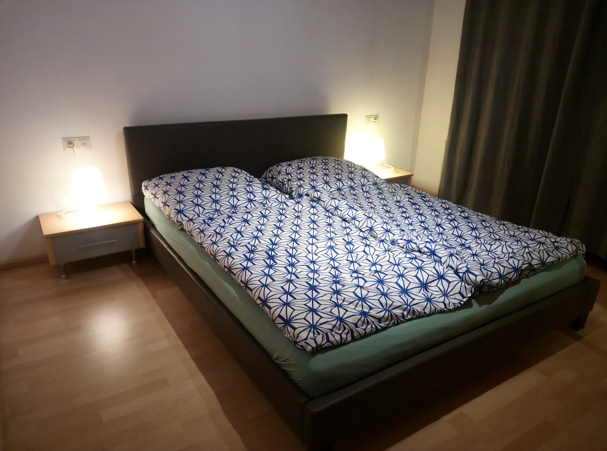A spacious bed is dressed with a patterned blue and white duvet cover, complemented by two bedside tables. Soft lighting is provided by lamps on each side, enhancing the calm ambiance of the sleeping area, with light wood flooring visible.