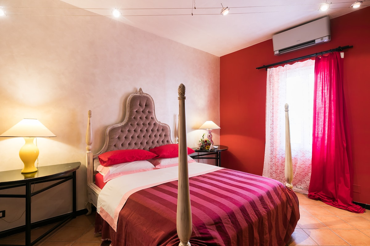 A well-appointed bedroom features a plush headboard and a spacious bed with red and white linens. Two bedside lamps provide lighting, complemented by vibrant red walls and curtains. A window allows natural light while a cozy atmosphere is maintained by the decor.