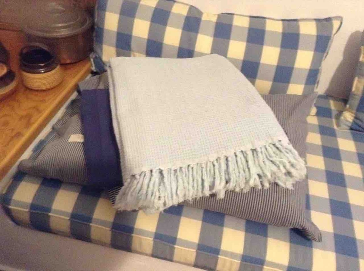 A cushioned seating area is displayed, featuring a combination of blue and white checkered, along with striped fabrics. Soft blankets in light blue and navy are neatly arranged on the seating, which is complemented by various round and rectangular pillows.
