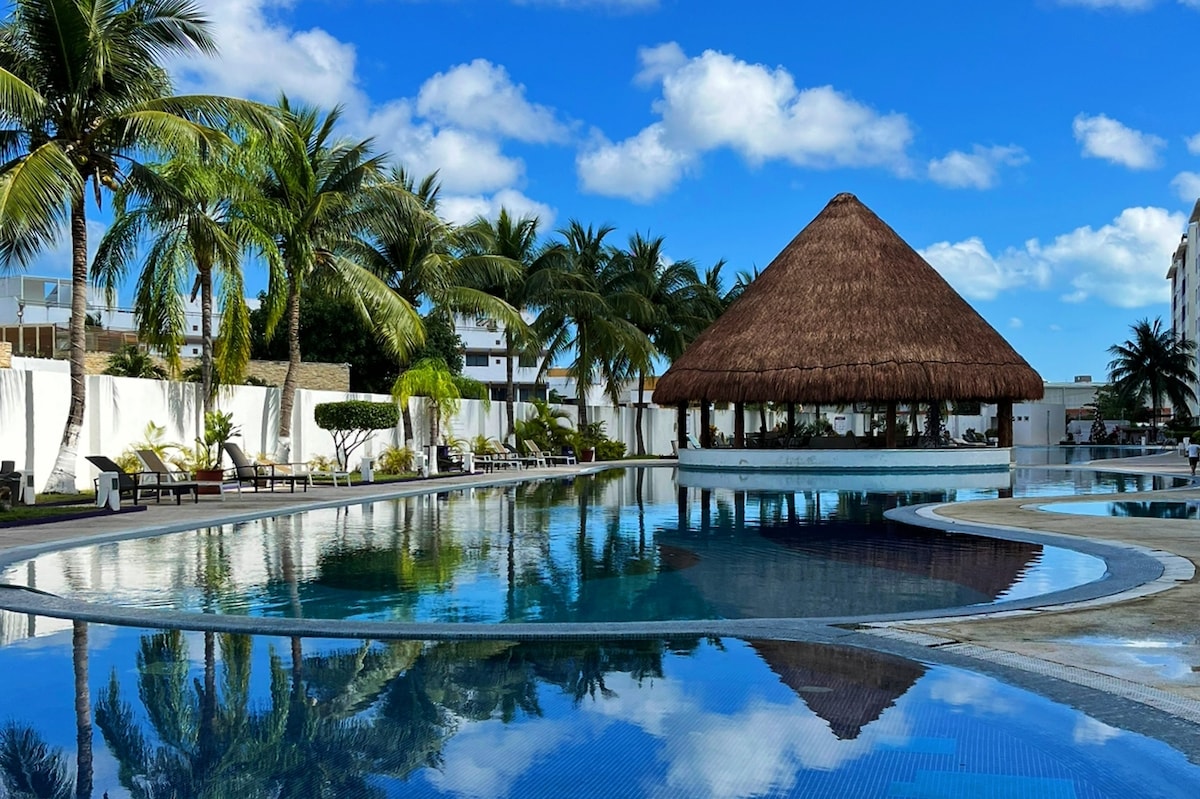 A large swimming pool reflects the clear blue sky and surrounding palm trees. A thatched gazebo provides shade, and sun loungers are placed around the pool area, creating a serene setting for relaxation.