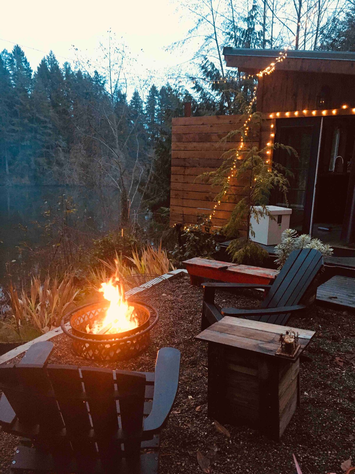 A fire pit sits surrounded by two black adirondack chairs and a small wooden table. Soft lights adorn the exterior of the tiny house, with trees and foliage providing a natural backdrop. The scene conveys a relaxing outdoor space near the water.