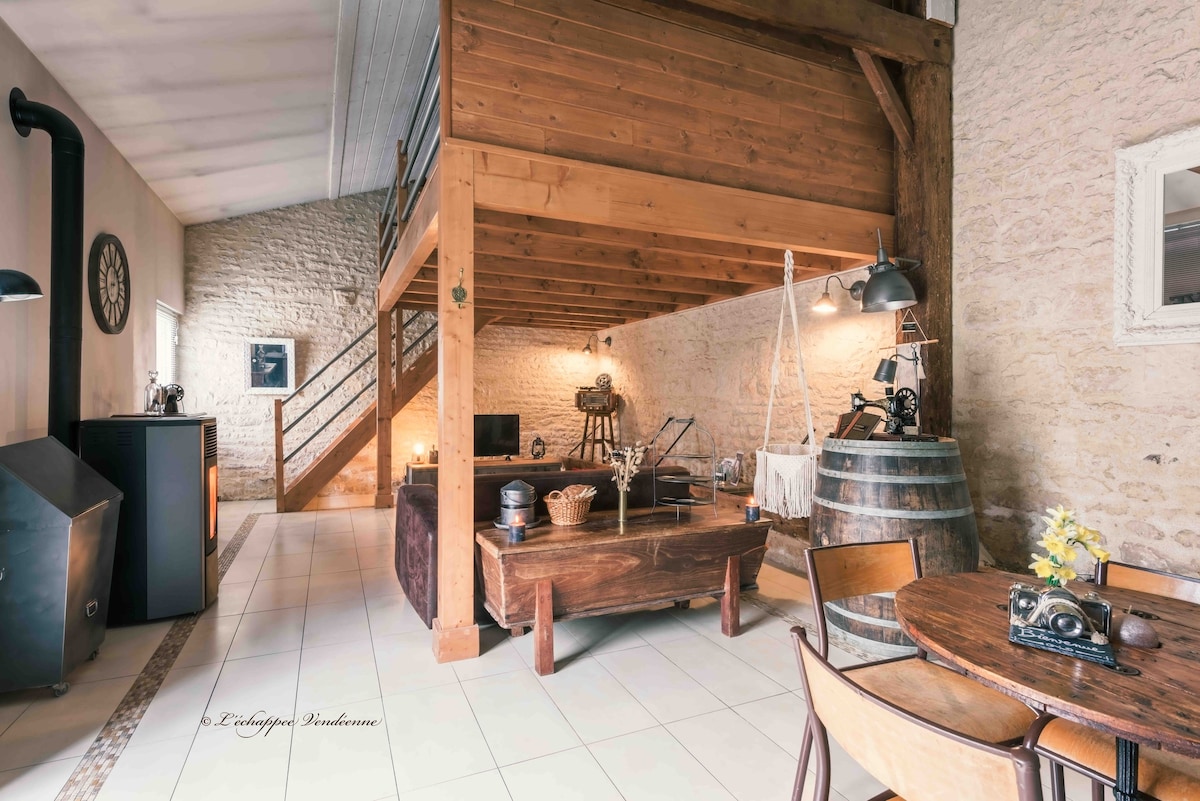 An open living space features a warm wooden loft above, with a cozy seating area on the lower level. Natural light filters through large windows, highlighting rustic elements like a wooden table, a decorative barrel, and a stylish fireplace.