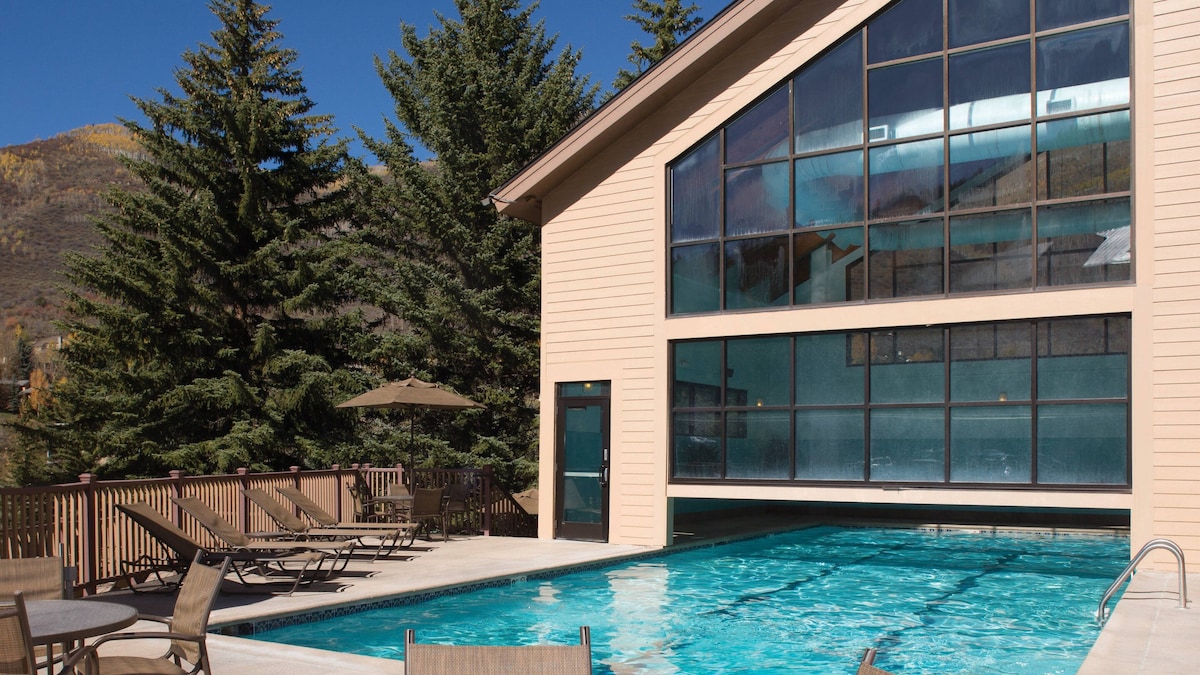 An outdoor pool area is framed by a large glass wall and surrounded by trees. Lounge chairs are positioned along the poolside, and a shaded area is provided by an umbrella. The clear blue water reflects the sky and surrounding landscape.