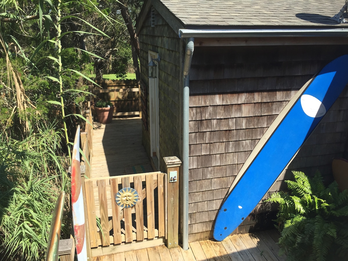 The image shows a wooden walkway leading to the entrance of the garage suite, bordered by lush greenery, including tall bamboo. A vibrant blue surfboard leans against the building, and a wooden gate marks the entrance to the private deck.