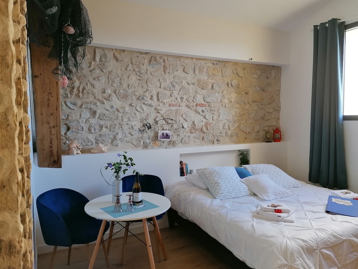 A cozy room features a spacious bed dressed with white linens and decorative pillows. A rustic stone wall adds character, while a small dining area hosts a round table with two blue chairs. Natural light streams in through a nearby window, enhancing the tranquil setting.