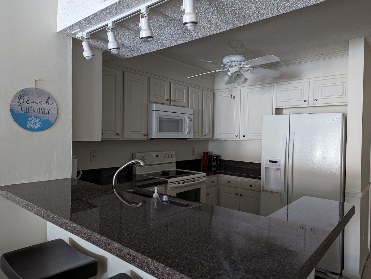 A compact kitchen features white cabinetry and dark granite countertops, providing ample workspace. Stainless steel appliances include a refrigerator, microwave, and oven. A ceiling fan above aids in air circulation, while modern lighting fixtures illuminate the cooking area. Bar stools line the counter, creating an inviting setup.