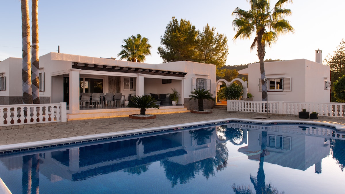 A contemporary garden villa is highlighted by a tranquil swimming pool, reflecting the clear sky. Palm trees and lush greenery surround the property, while a spacious terrace with dining seating is visible, promoting a relaxed outdoor atmosphere.