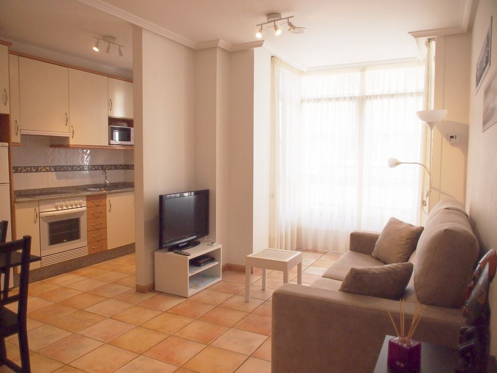 A well-lit living area showcases a comfortable sofa and a coffee table, with a television positioned on a white stand. The adjacent kitchen features contemporary cabinetry and essential appliances. Large windows allow natural light to fill the space, enhancing the room's openness.