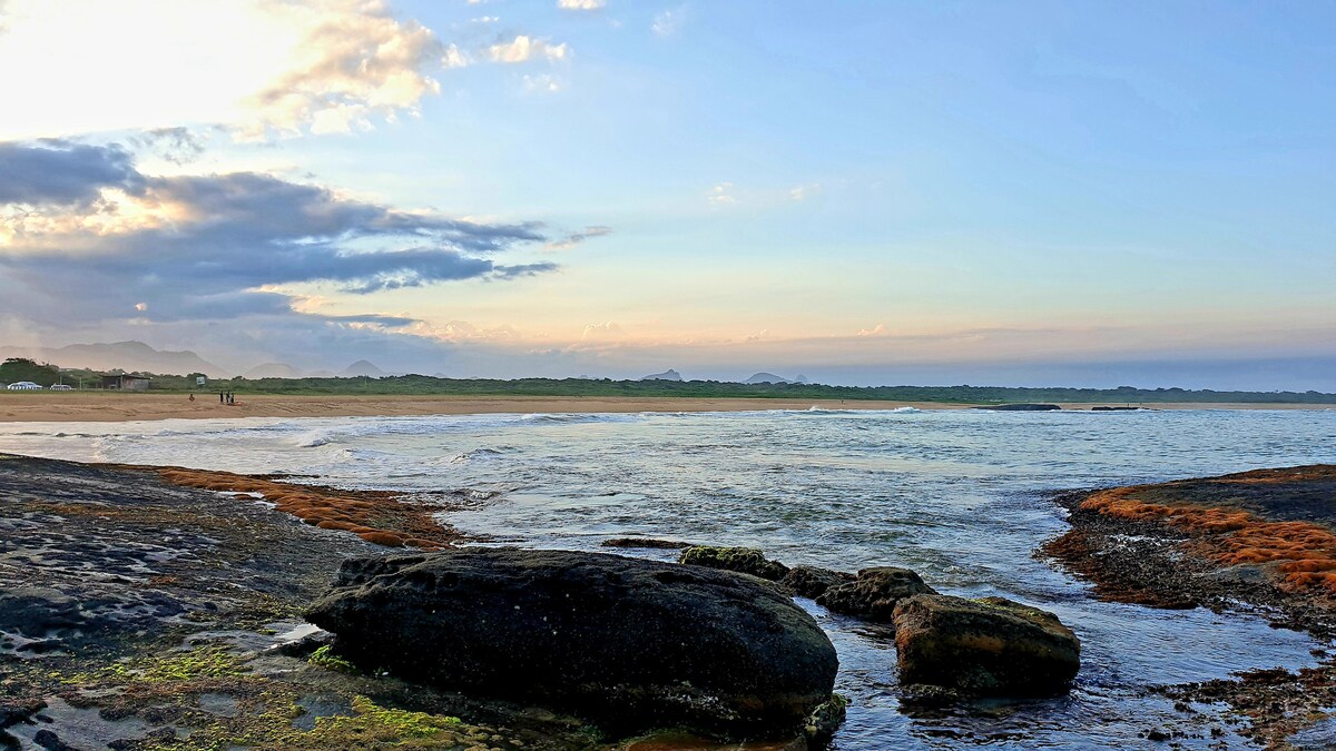 An expansive beach scene captures the shoreline where water meets sand and smooth rocks. Gentle waves lap against the shore, and soft clouds hover in the sky, reflecting shades of blue and golden light. The distant landscape features hints of greenery and sandy stretches.