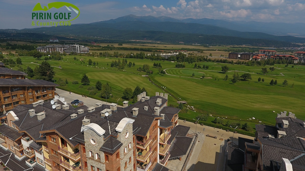 A panoramic view reveals the expansive layout of Pirin Golf & Country Club, featuring well-tended green golf courses framed by mountains in the background. Multiple residential buildings outline the complex, showcasing charming architectural details under a clear blue sky.