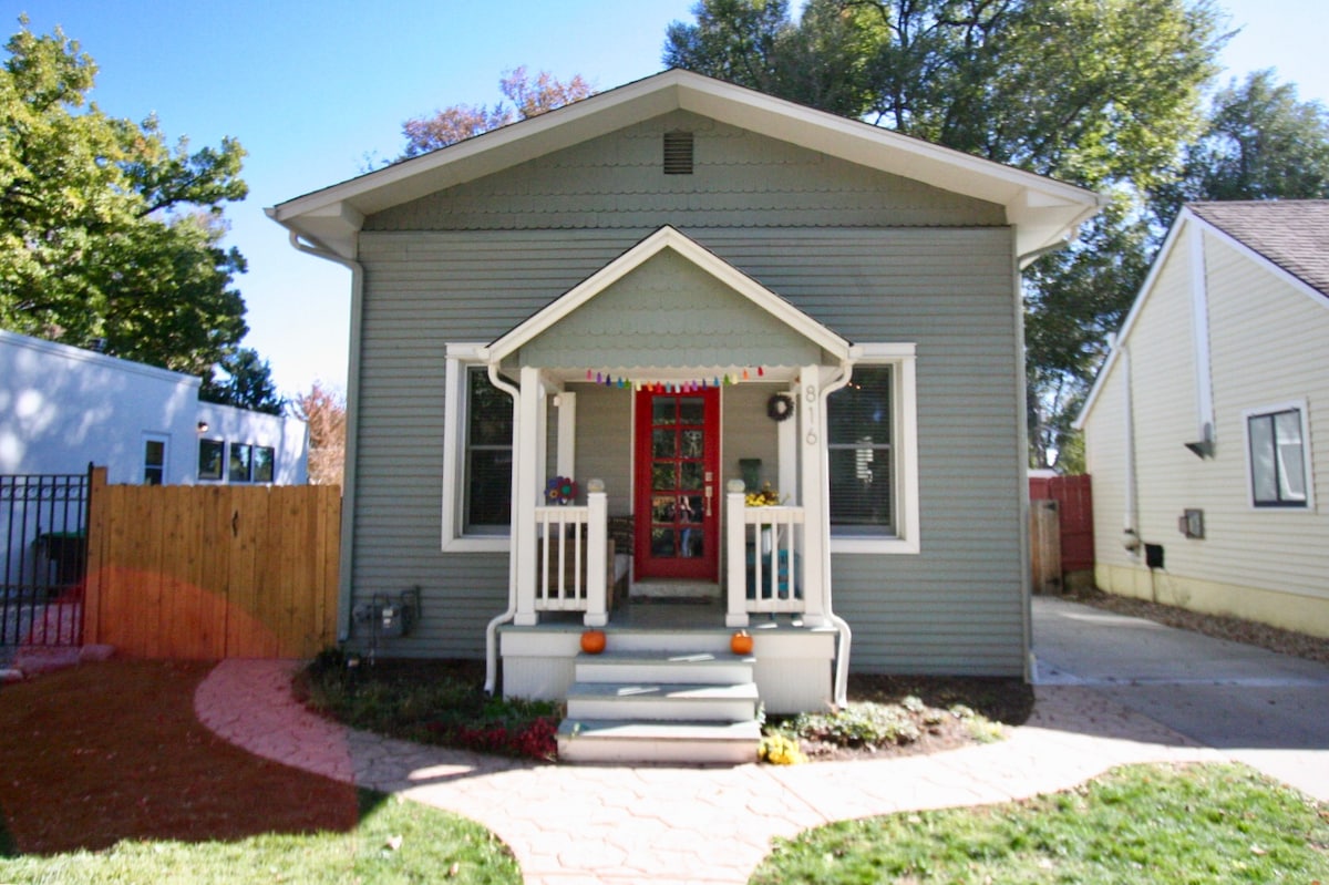The exterior of a modern grey home is showcased, featuring a welcoming front porch with white railings. A bright red door serves as the focal point. Seasonal decorations and a small garden with colorful flowers and pumpkins are present, while a paved pathway leads to the entrance.