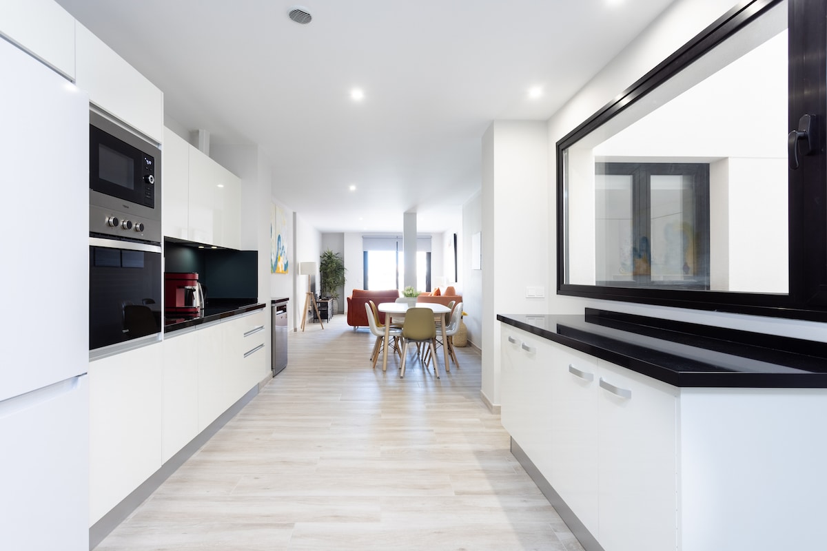 A modern kitchen features sleek white cabinetry and a black countertop, with built-in appliances, including an oven and microwave. Natural light filters through a large window, illuminating the spacious layout that connects to a dining area in the background.