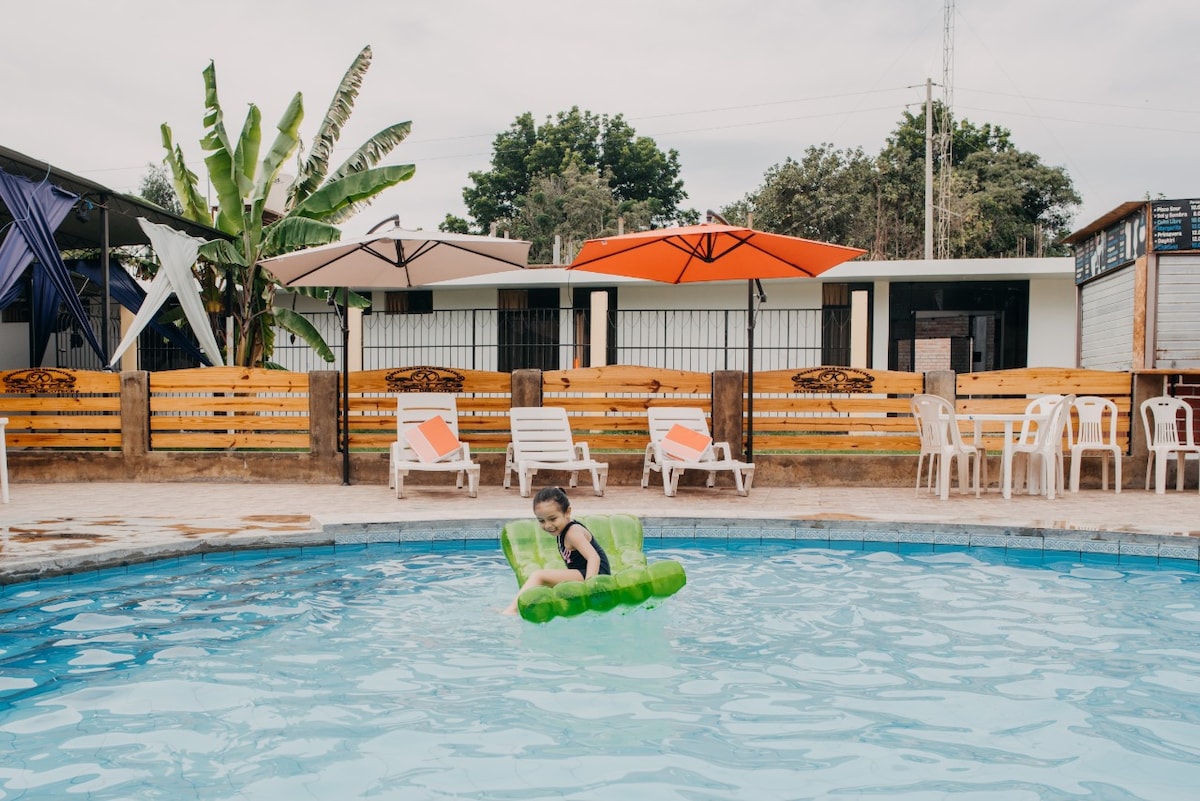A vibrant swimming pool is showcased with a person relaxing on a green float. Surrounding the pool, lounge chairs and umbrellas provide shaded seating. In the background, colorful umbrellas are seen above a well-maintained area, enhancing the inviting atmosphere of the outdoor space.