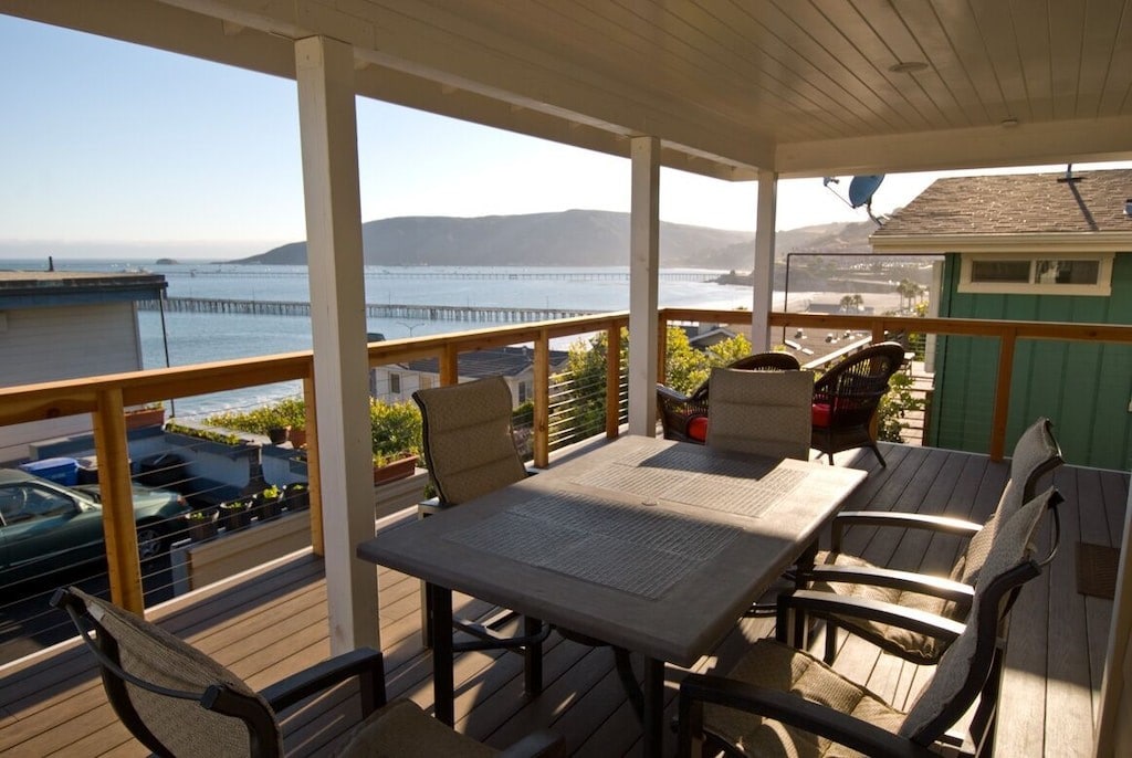 A spacious covered deck features a large outdoor table surrounded by six chairs, offering views of the ocean and surrounding hills. The deck's rail provides safety while enhancing the panoramic scenery of Avila Beach and its pier.