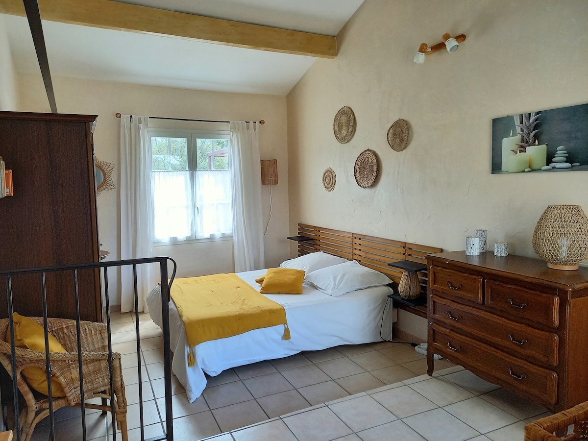 A spacious bedroom features a comfortable bed adorned with a yellow throw pillow. Natural light filters through sheer curtains, illuminating the room. Decorative wall art and woven baskets add texture, while a wooden dresser and a chair create a welcoming ambiance.