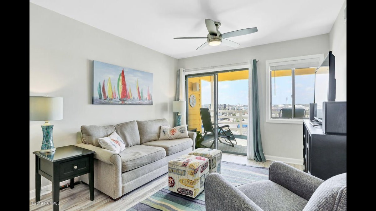 The living area is designed for comfort, featuring a plush sofa and an ottoman with a colorful pattern. A ceiling fan can be seen above, and large windows offer bright natural light and views. A vibrant painting of sailboats complements the decor.