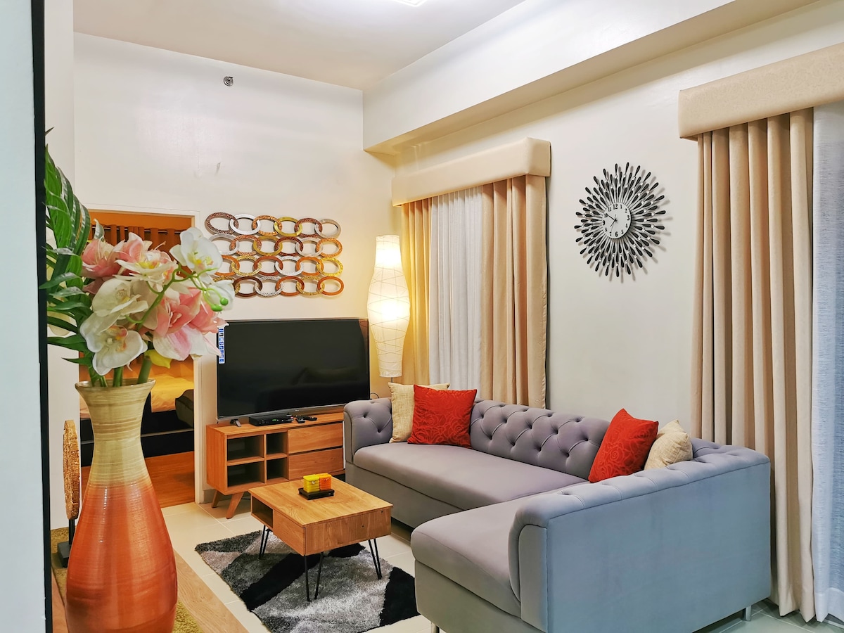 The living area features a modern L-shaped sofa with decorative throw pillows, a wooden coffee table, and a large smart TV. Soft lighting is provided by a floor lamp, and natural light enters through sheer curtains. A decorative wall hanging adds visual interest to the space.