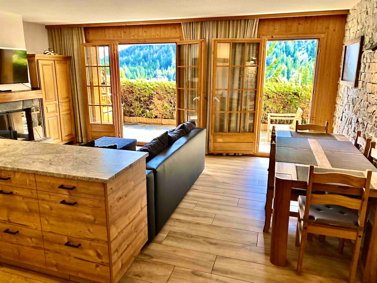 An open living area features large glass doors with scenic mountain views. A cozy sofa faces a stone fireplace, while a wooden dining table is positioned nearby. Ample natural light enhances the warm wood tones throughout the space.