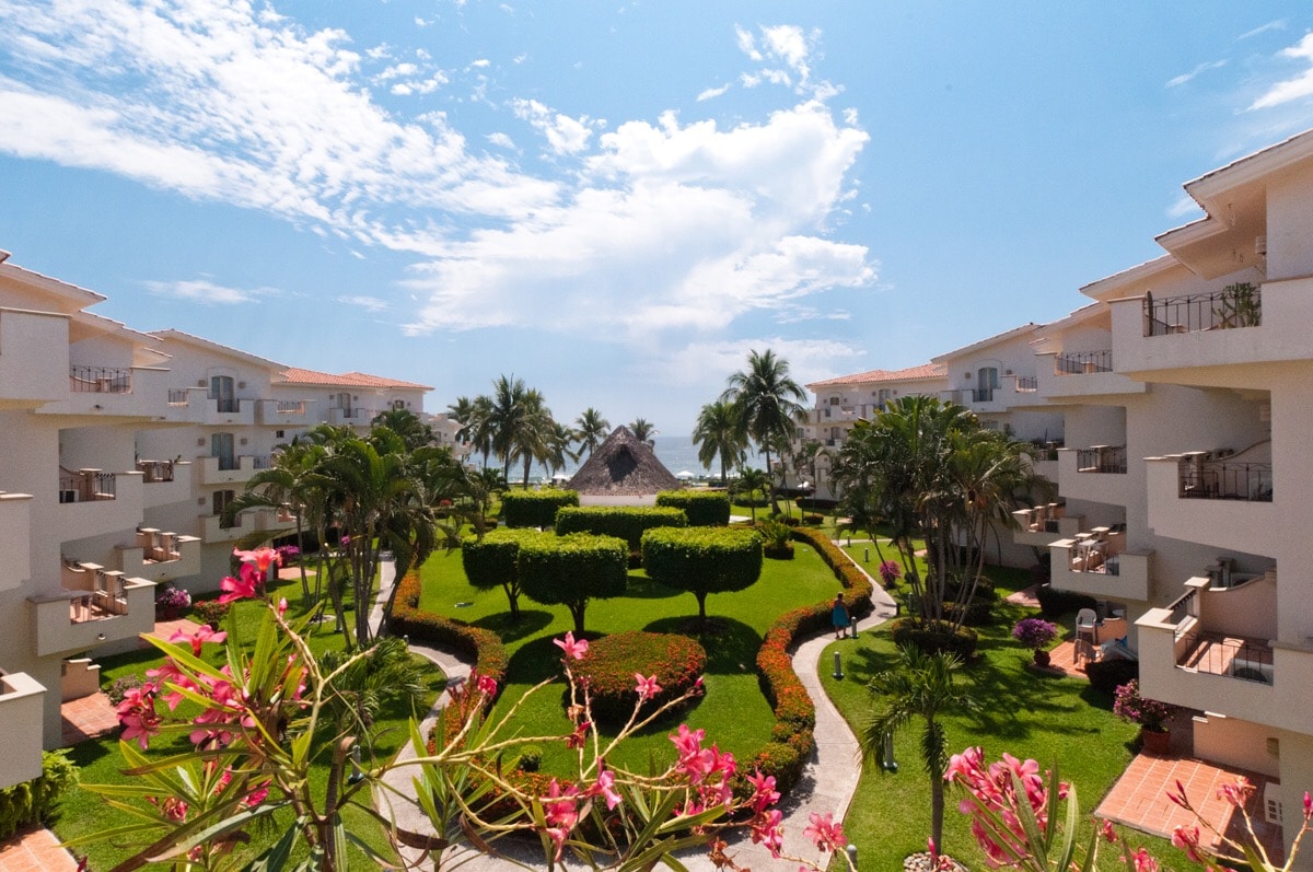 The image captures a vibrant landscape within the condo complex, featuring well-maintained gardens bordered by palm trees. A traditional palapa stands in the background, while ocean views are visible beyond the lush greenery. The setting offers a sense of tranquility and access to nature.