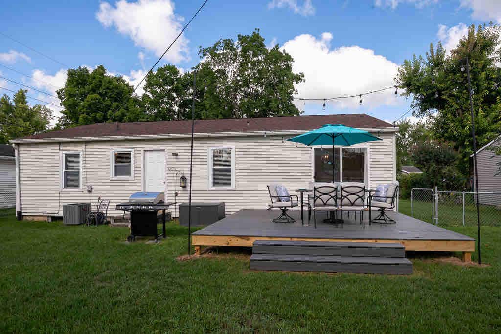 A well-maintained backyard features a wooden deck with seating and an umbrella, surrounded by a lush lawn. A gas grill is positioned on one side, while the house displays an inviting facade with multiple windows and a door, framed by green trees.