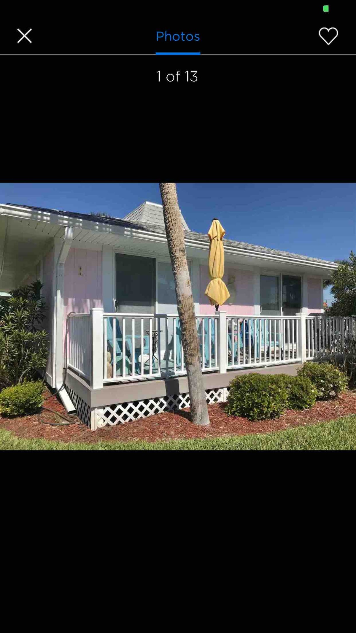 The exterior of the beach cottage features a pale pink facade, accented by white railings and a sunny yellow umbrella. A small porch is equipped with several blue chairs, surrounded by well-kept shrubs and greenery, enhancing the inviting appearance of the property.