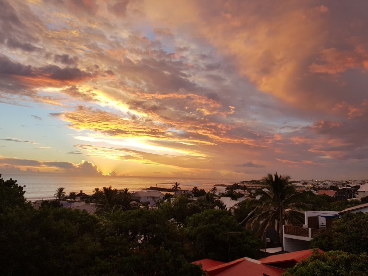 A vibrant sunset fills the sky with warm hues of orange and purple, overlooking a coastal view. Palm trees frame the scene, while the tranquil ocean glistens in the fading light. The rooftops of nearby buildings are visible, creating a serene atmosphere.