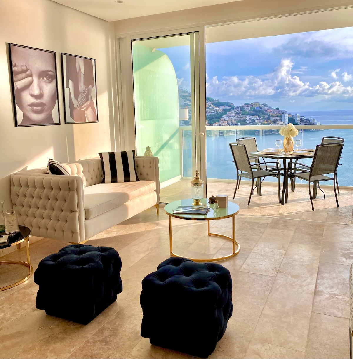 A stylish living area features a comfortable seating arrangement with a plush sofa and decorative cushions. A glass coffee table sits in front, complemented by two upholstered ottomans. Floor-to-ceiling windows offer expansive views of the coastline and bay, enhancing the bright ambiance.