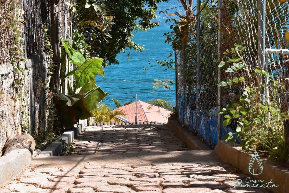 A stone pathway leads downward, flanked by greenery on either side. The path descends towards a tranquil body of water, visible in the distance, with a rooftop peeking through the trees. A gentle slope indicates the approach to the lake.