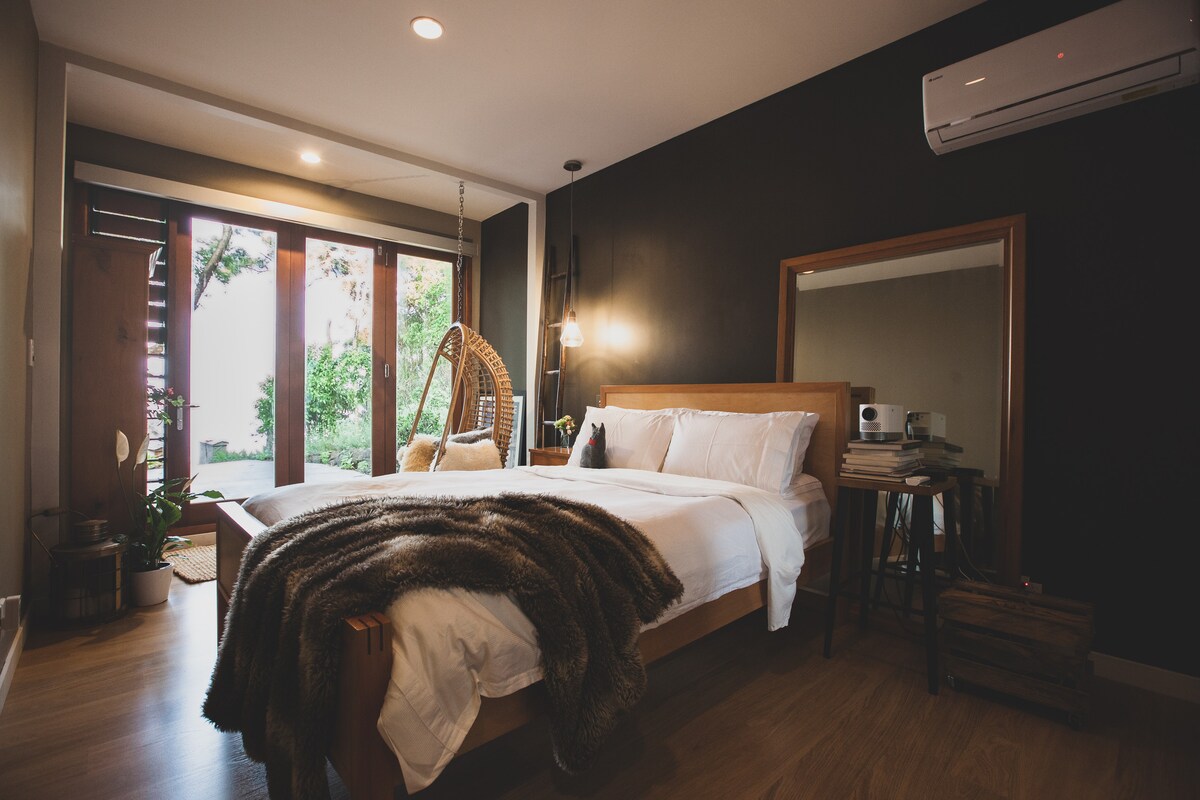 A private room features a spacious wooden bed covered with a soft blanket. A large mirror and a plant are placed beside the bed. Natural light floods in through sliding glass doors, which open to a green outdoor space. A hanging chair and air conditioning unit enhance comfort.