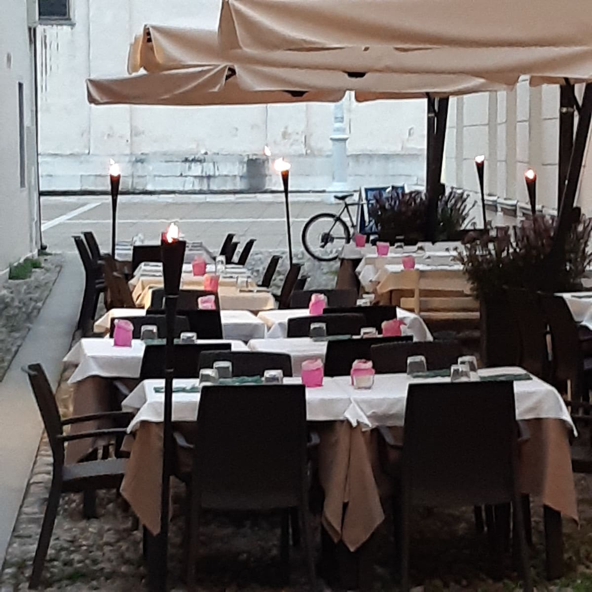 An outdoor dining area is shown, featuring multiple tables set with white tablecloths and decorative centerpieces. Torch-lit ambiance is provided by flames on tall stands. A bicycle is visible in the background, alongside the building's facade. The overall atmosphere appears inviting for diners.