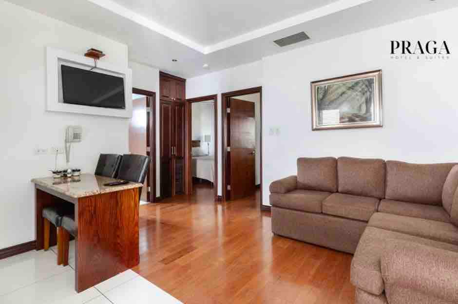 A spacious living area is shown, featuring a comfortable beige sofa and a wooden dining table with two chairs. A flat-screen TV is mounted on the wall, and doors lead to the adjoining rooms. Natural light enters through the windows, highlighting the wooden flooring.